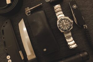 Black Essentials – A Masculine Assembly of Everyday Carry Items A meticulously arranged tableau of black EDC (Everyday Carry) items lies on a graphite-gray cloth: a minimalist watch with a matte case, a sleek leather wallet, a Japanese tanto-style knife, a Swiss Army knife, a braided keychain, and a compact flashlight. Each object is positioned with intentional precision, their monochrome tones amplifying textures—brushed metal, pebbled leather, and polished steel. A study in utilitarian elegance, where function meets silent sophistication.
