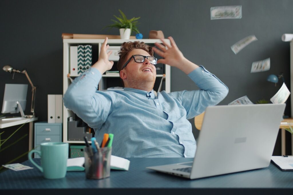 Entrepreneur attractive guy is relaxing in chair in office while money banknotes are falling down from above. Happy young people, wealth and business concept.